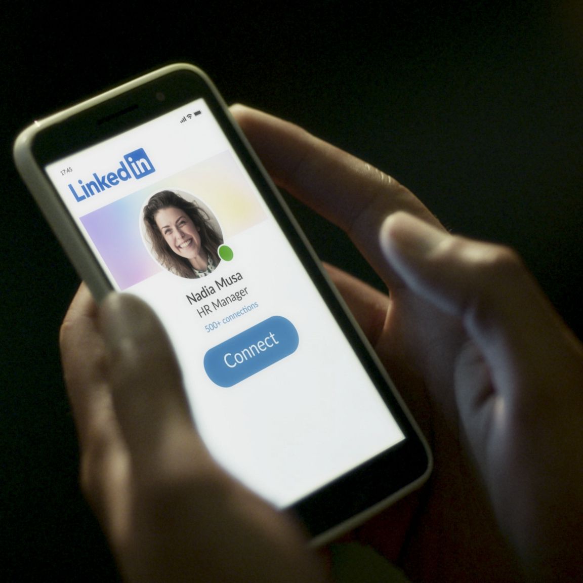 Hands holding a phone displaying the LinkedIn profile and photo of a human resources manager.