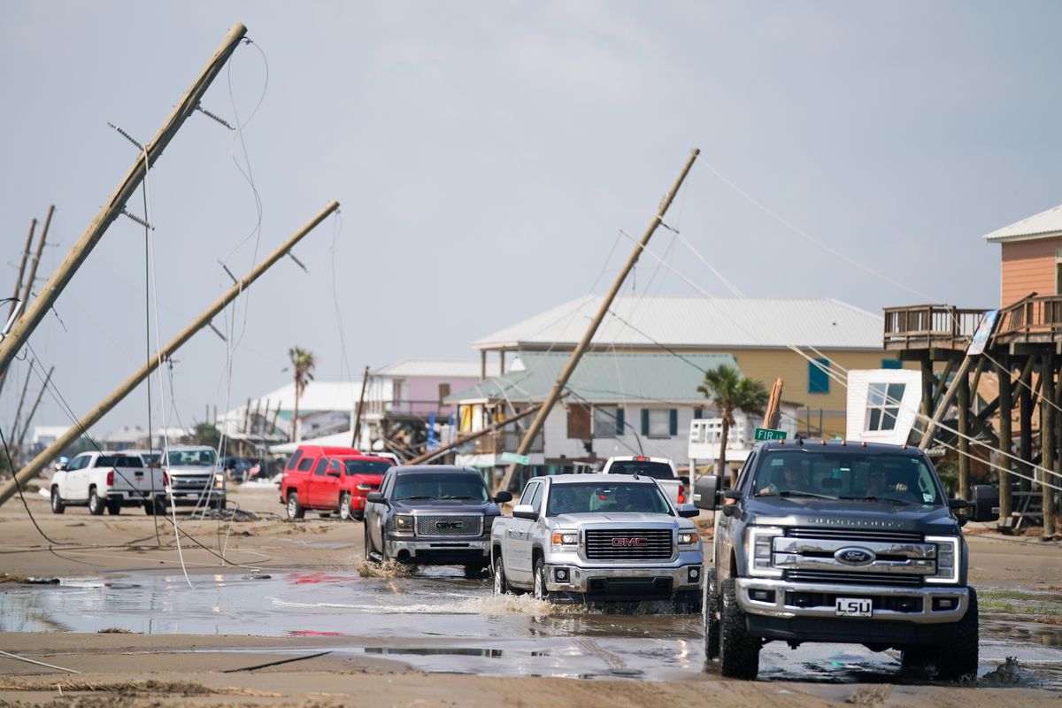 A line of pickup trucks drives through a muddy puddle in Grand Isle, Louisiana. Power poles are tilted at odd angles in the background, their cables falling on the ground and into the water.