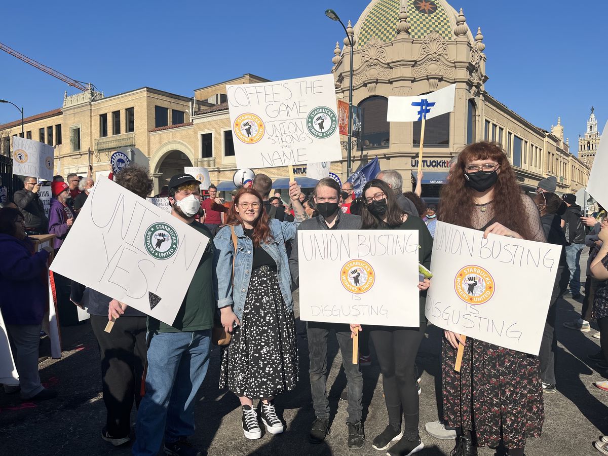 Starbucks workers stand in the street holding signs that read, “Union yes,” “Union busting is disgusting,” and “Coffee’s the game, union strong’s the name.”