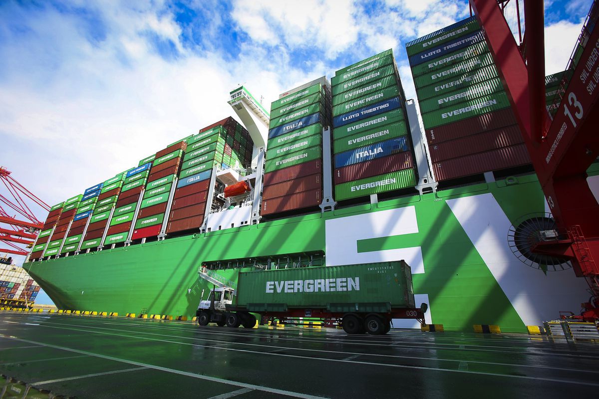 A huge container ship docked, with a truck hauling a container beside it.