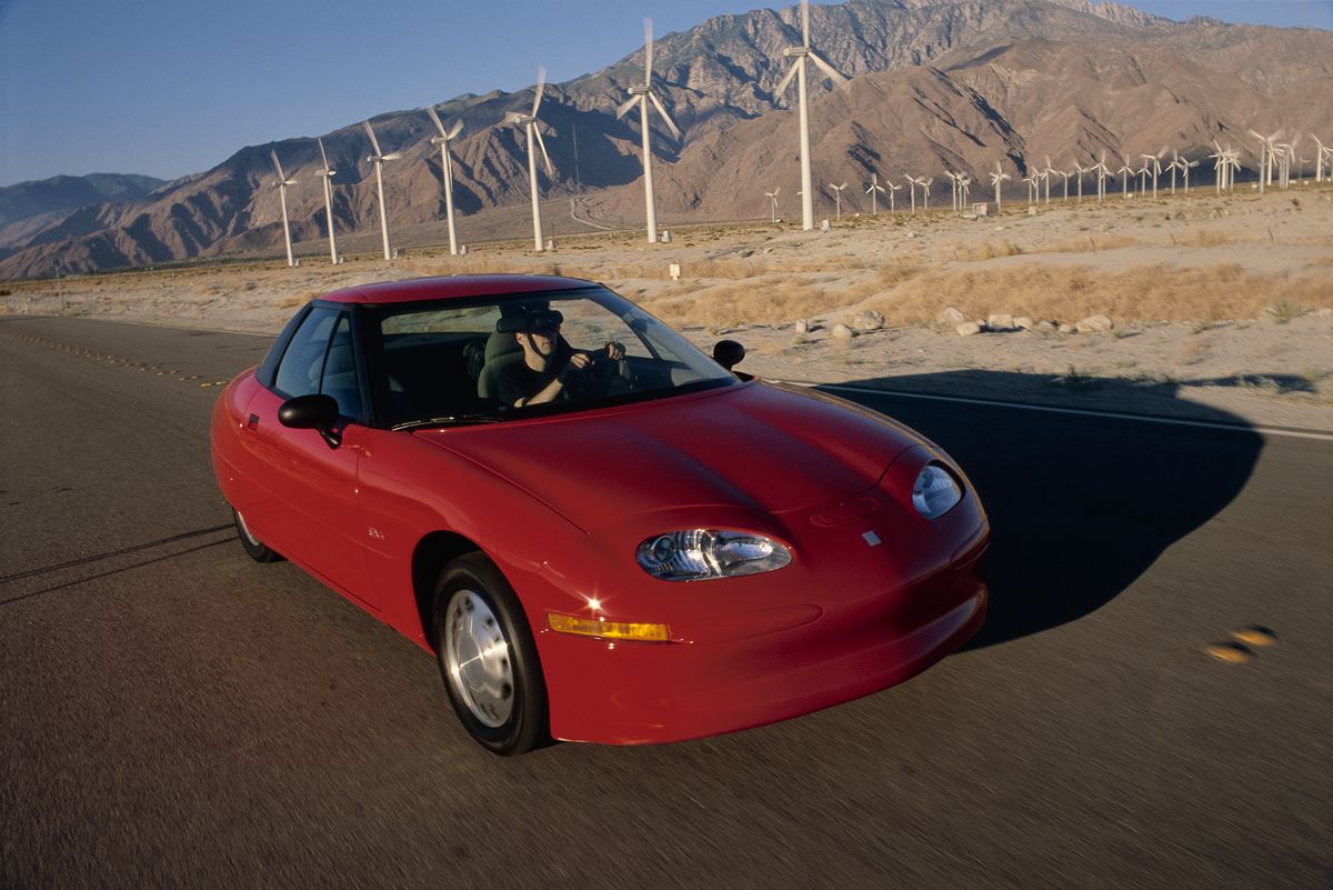 A small red electric car being driven past a wind farm.