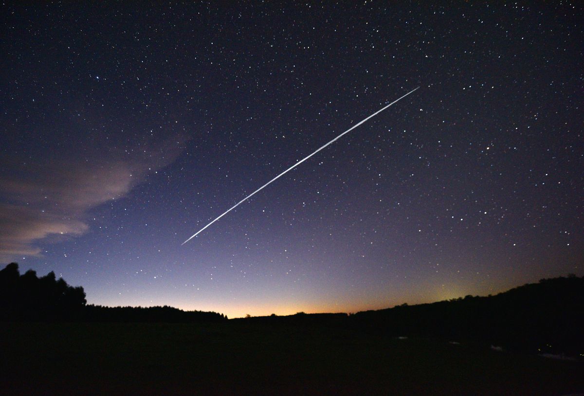 A streak of light across a dark star-filled sky as light appears over the horizon.