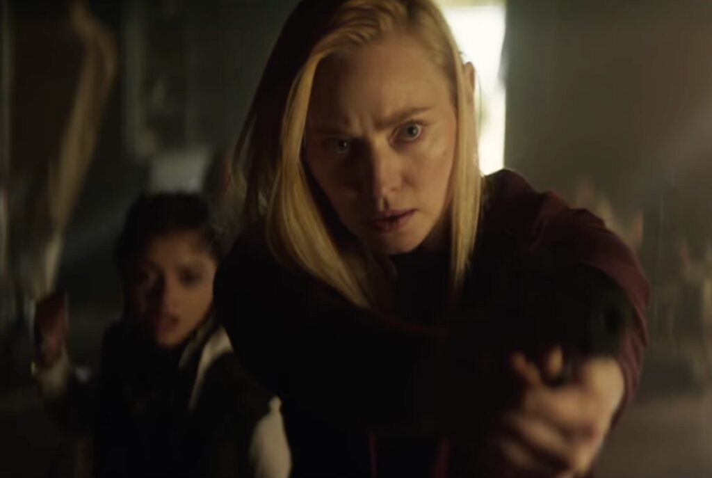 blonde woman with determined focused expression pointing a gun at something in front of her while shielding a little girl behind her
