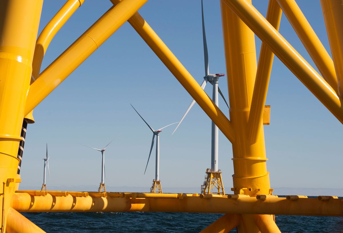 Four large, white offshore wind turbines are seen stationed in ocean water through the yellow support struts of a fifth turbine.