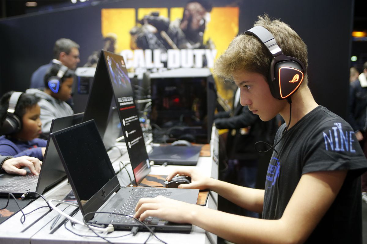 A young man wearing large headphones sits at a laptop at a table with other people, each playing a video game.