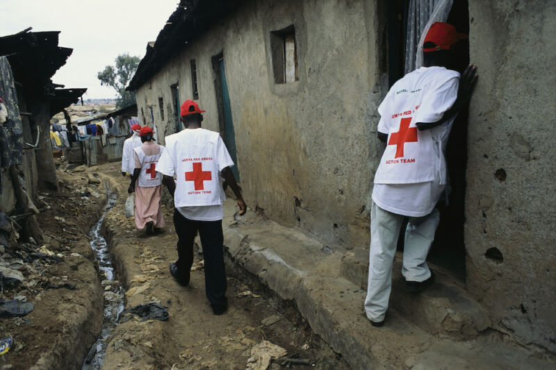 People in Red Cross vests walk along a dirt street.