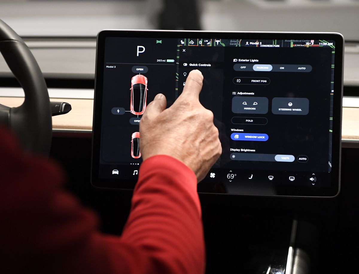 A driver with their hand touching the Tesla screen beside the car’s steering wheel.