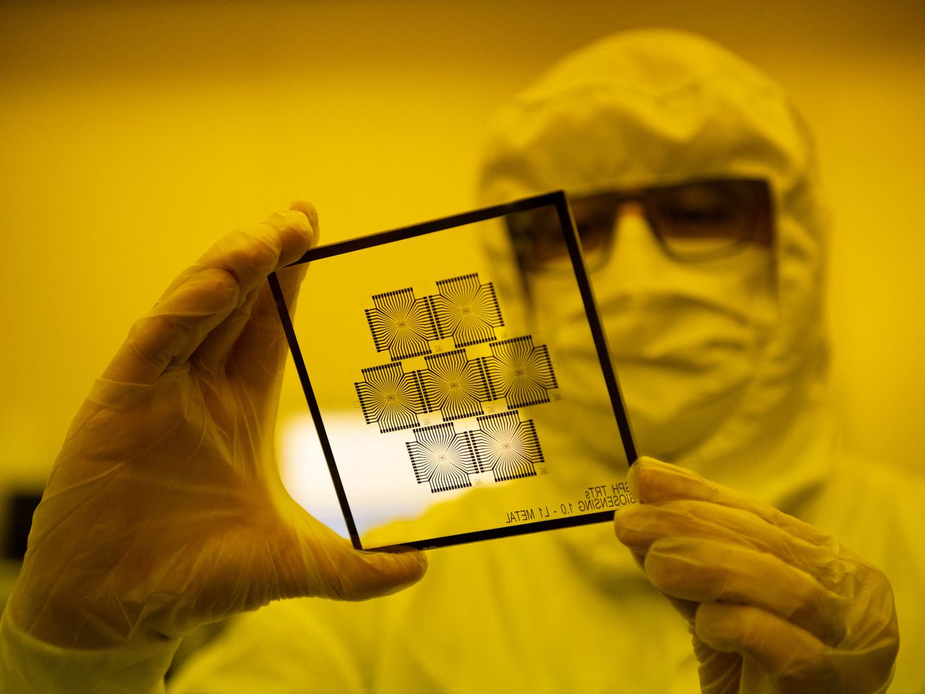 An employee working in a chip manufacturing clean room of the Barcelona Institute for Microelectronics holds up a glass pane to the light.