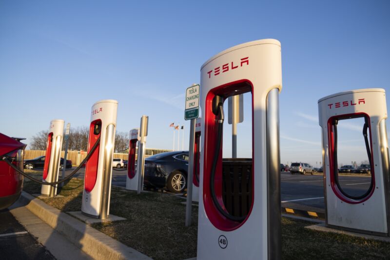 A half-dozen Tesla chargers in a parking area, with a couple Tesla vehicles getting a charge.